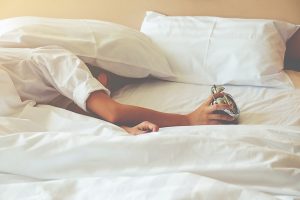 A person in bed with a pillow over their head holds an alarm clock